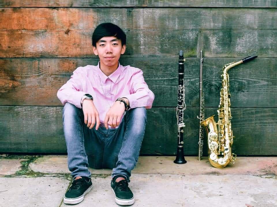 photo of composer Jonavin Thao siting on the ground next to an alto sax, a flute and a clarinet